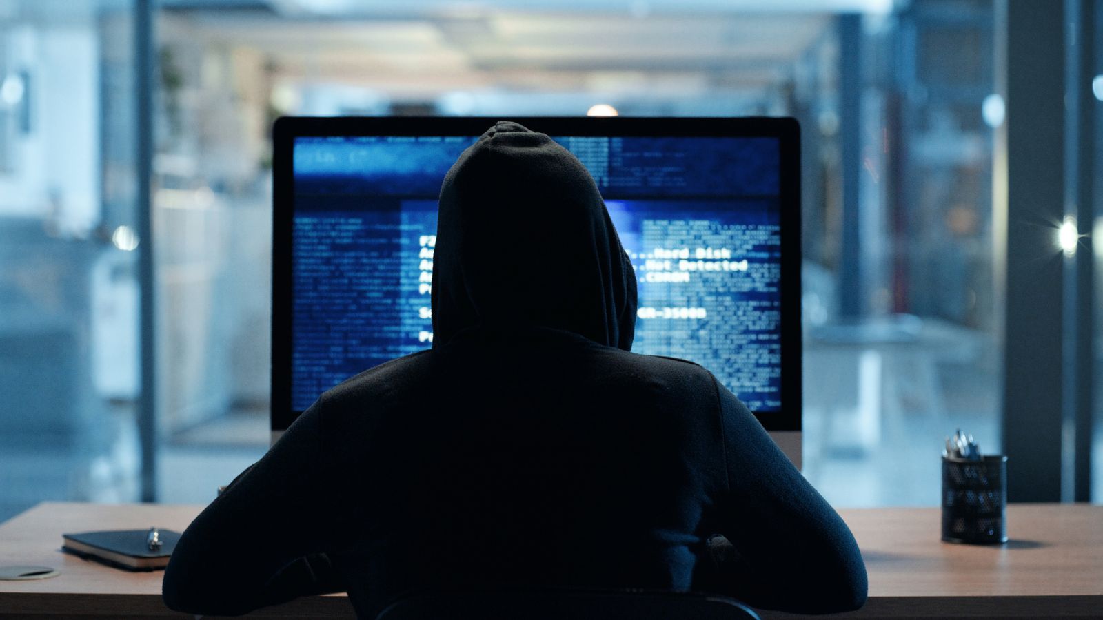 Back of hacker or cyber criminal stealing information online sitting at a computer at night. Hooded mysterious person coding spyware, malware and viruses to commit fraud, theft and phishing scam