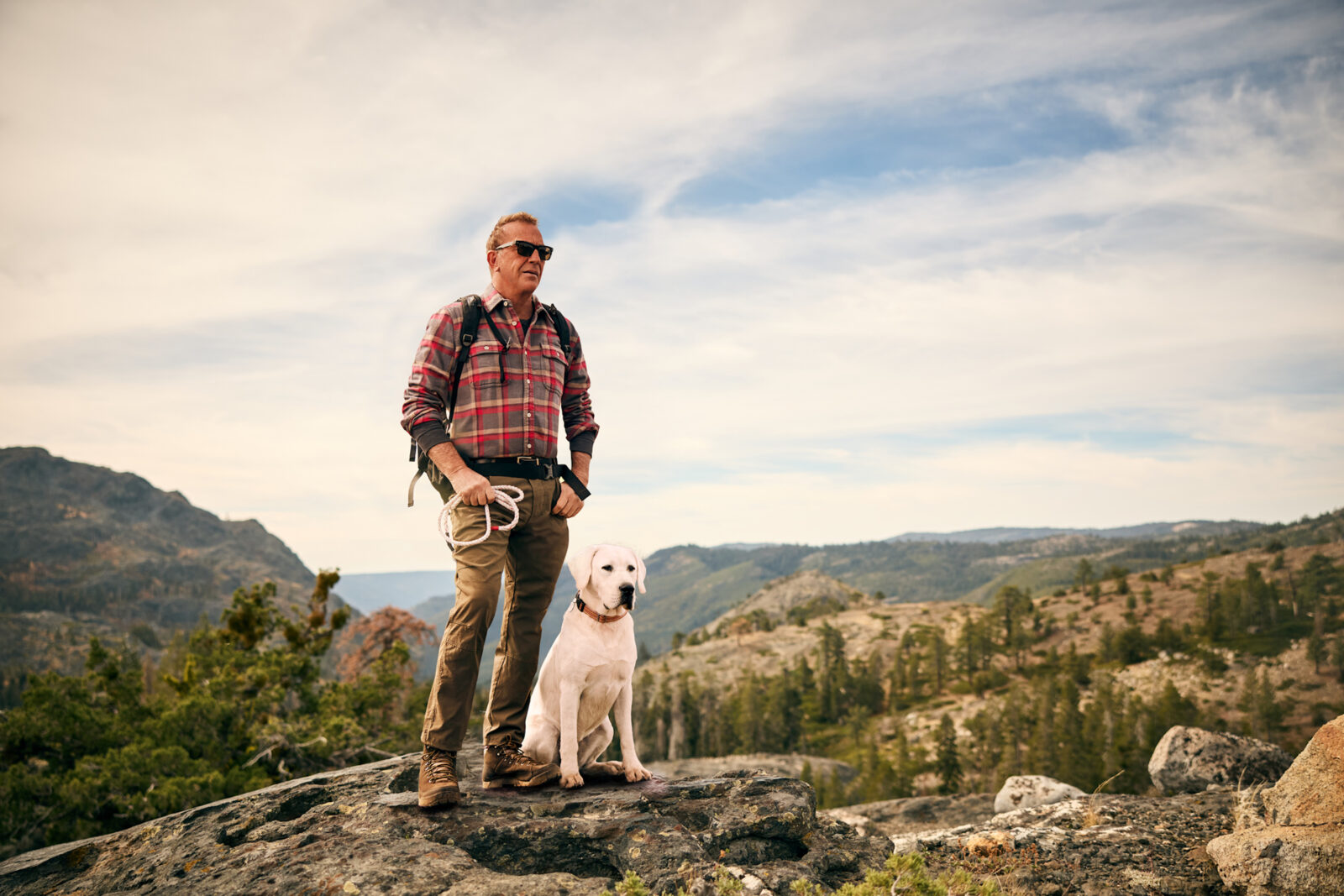 A man and dog stand on a rock against a mountain landscape